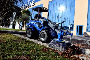mini loader with a side brush attached, cleaning leaves in front of multione venue