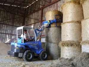 man driving a mini loader and using an hay bale grapple to grab a hay bale, seen from behind